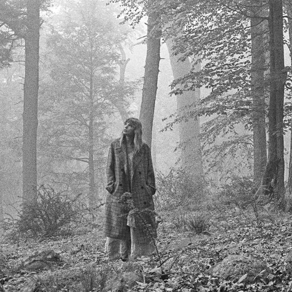 A woman (Taylor Swift) stands amidst tall trees in a foggy forest, surrounded by a mysterious, ethereal atmosphere.