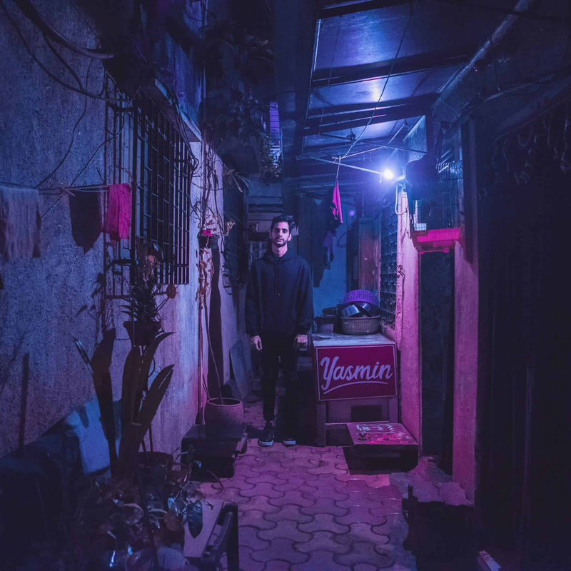 A person stands in a dimly-lit, narrow alley with plants and a sign reading "Yasmin."