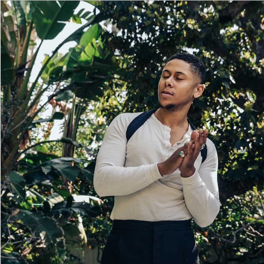 A person in a white shirt with suspenders standing among green foliage checking their hands.