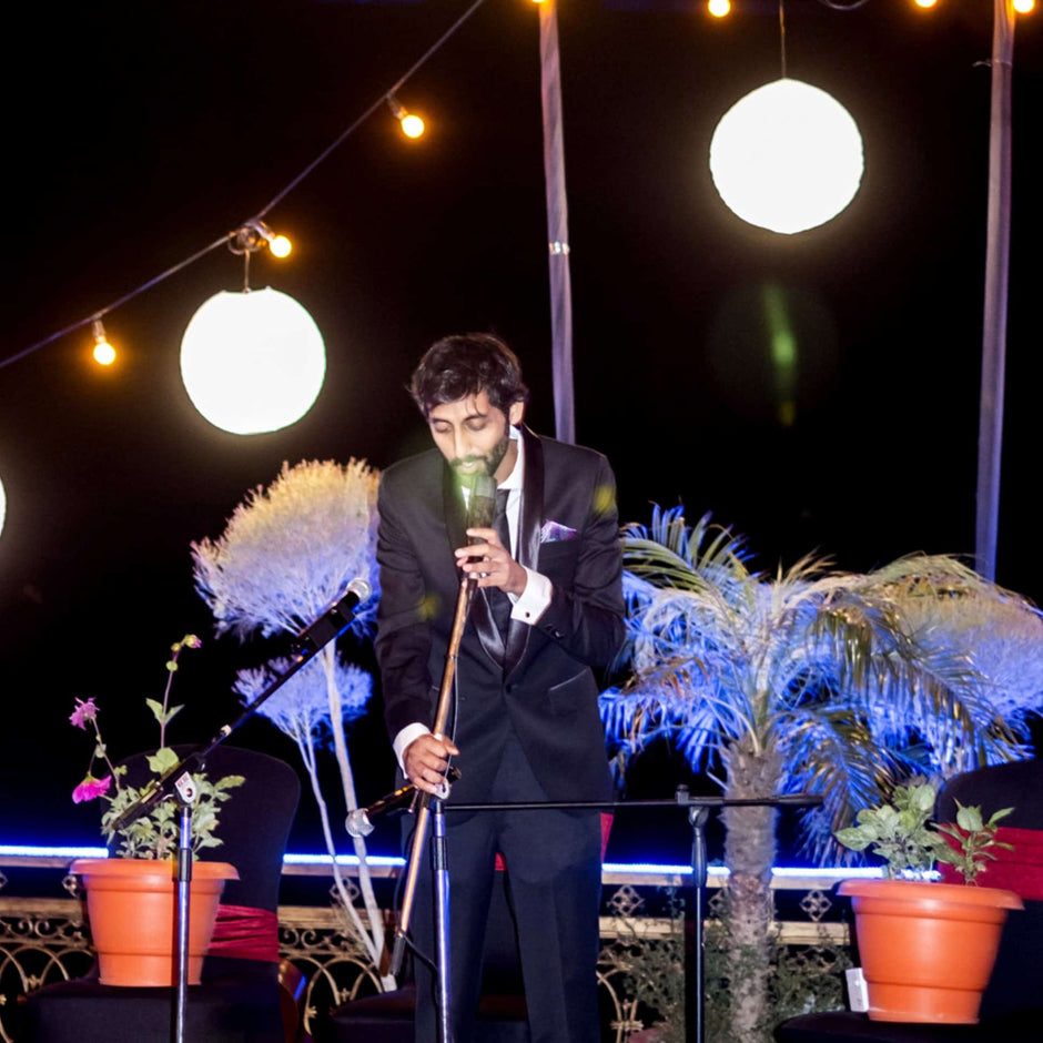 Person in a suit singing in a microphone at an outdoor nighttime event with string lights.