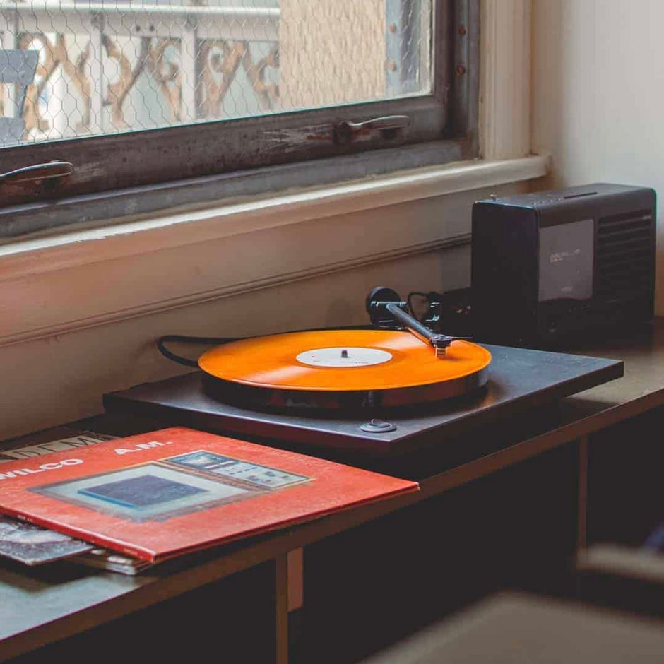 A turntable with a vinyl record playing in it and the album art on the side placed on a table near a window 