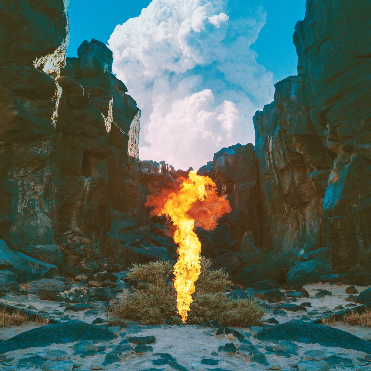 A vibrant fire blazes in the center of a vast, arid desert landscape under a blue cloudy sky.